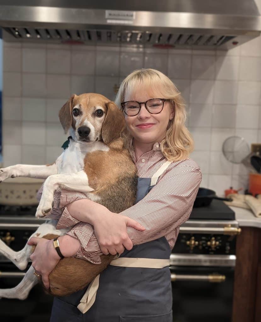 Sam in a striped shirt and blue apron holding her beagle, Hesh.