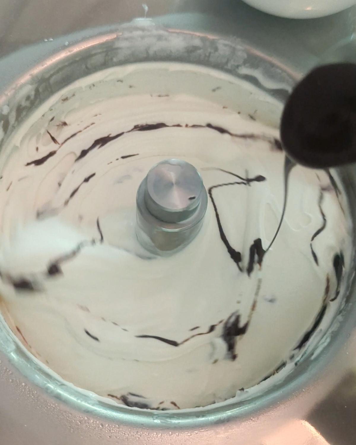 overhead view of a spoon drizzling stracciatella chocolate into a batch of ice cream in a running compressor machine.