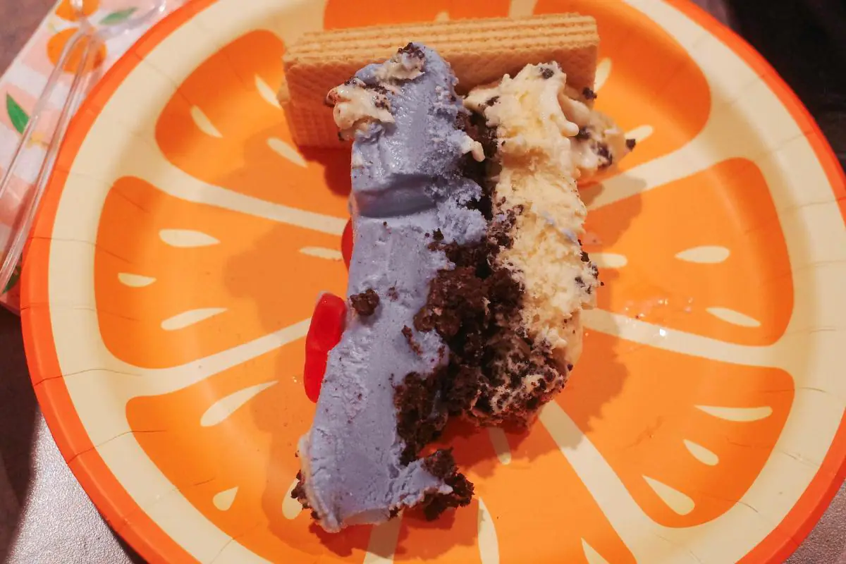 a slice of swimming pool ice cream cake on an orange plate showing the layers of blue ice cream, ice cream cake crunchies, and vermonty python ice cream.